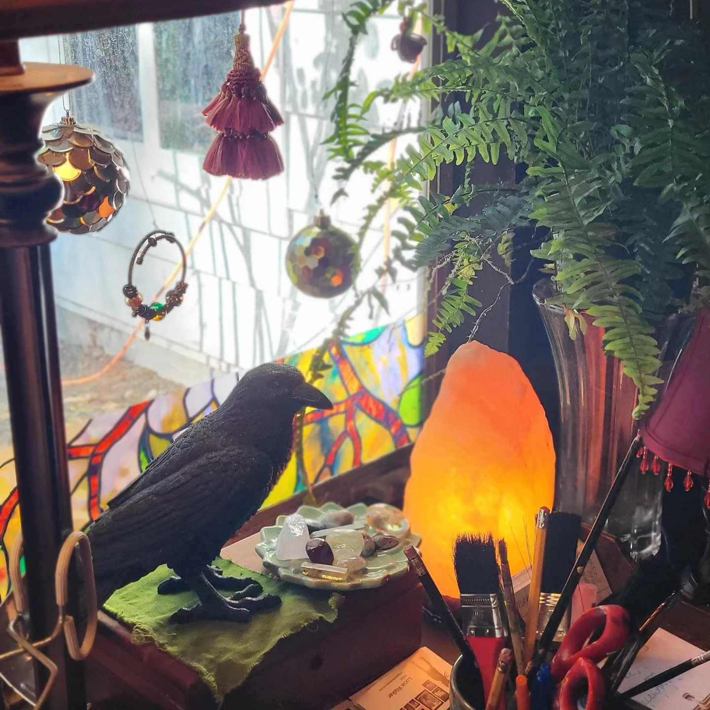 photo by Lucie Walker photof of a ceramic crow, a ceramic plate of gemstones, a lit salt lamp, a fern, some hanging ornaments in a window